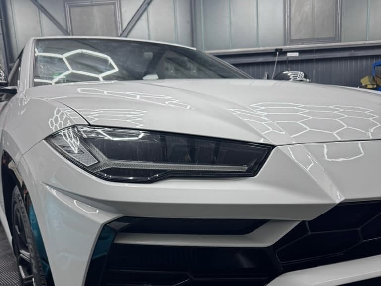 Front eines weißen Lamborghini Urus mit glänzendem Lack, in dem sich die Werkstattbeleuchtung spiegelt
